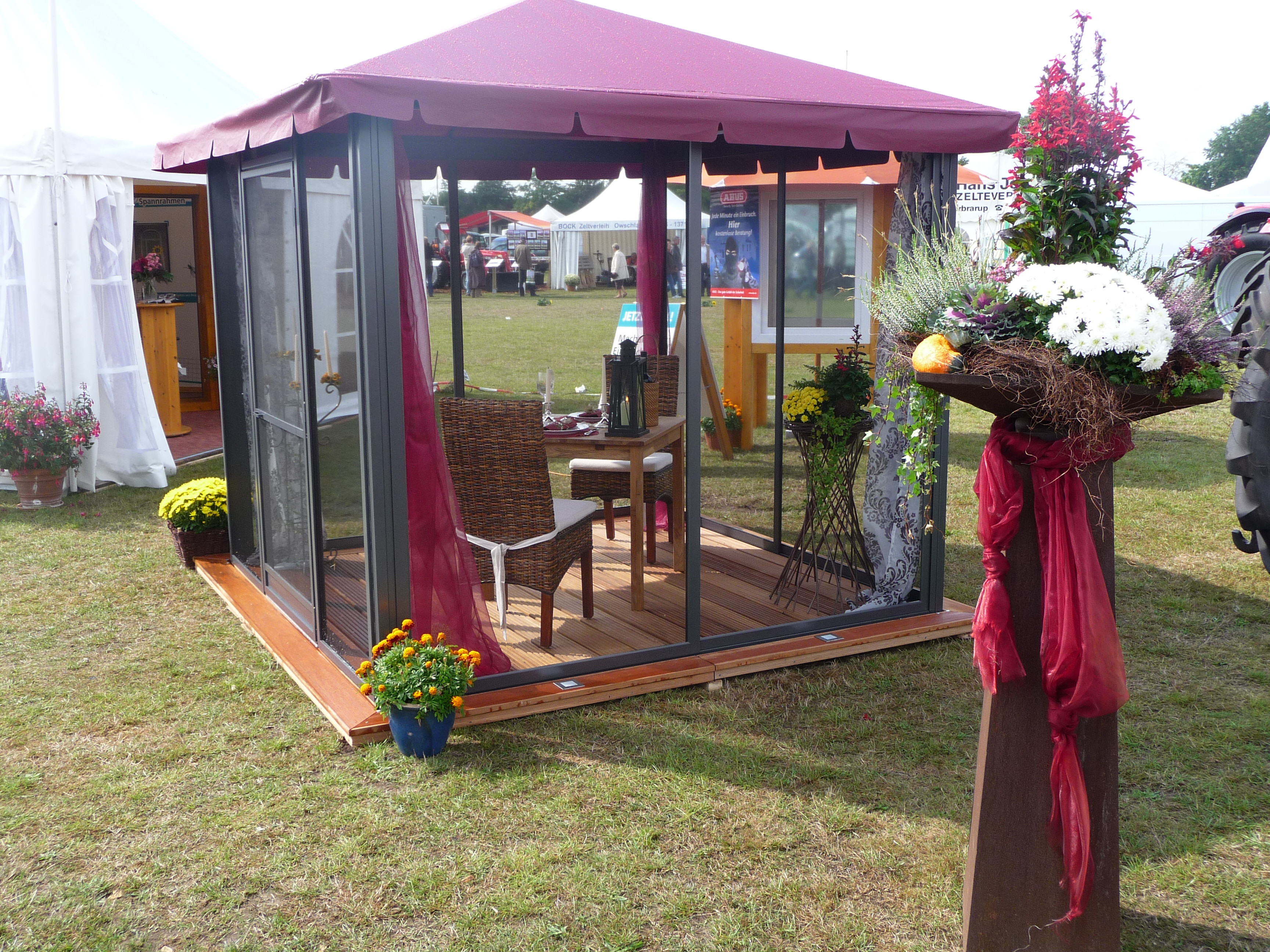 Ein Pavillon mit Fliegengittern auf allen 4 Seiten. Im Inneren kann man mit 2 Personen unter dem roten Dach sitzen. Um den Pavillon stehen unzählige Blumen, die bei Sonnenschein zum sitzen einladen. Schmidt Insektenschutzrahmen GmbH